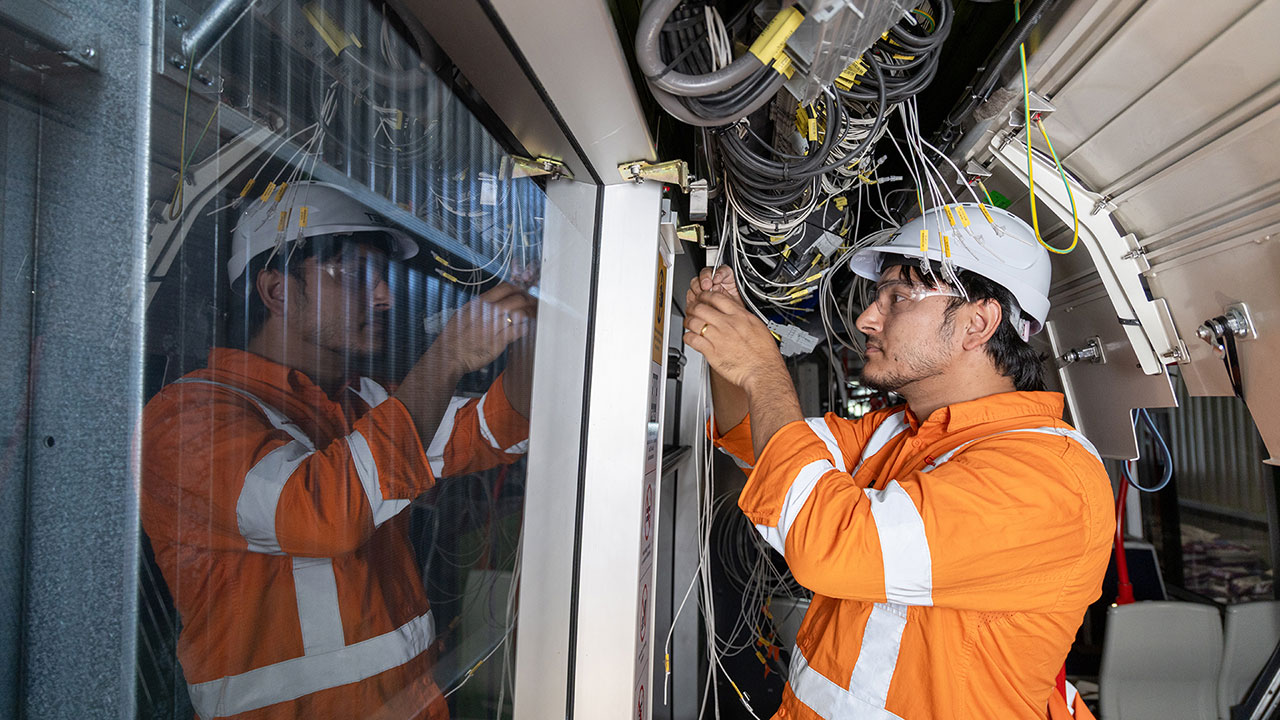 A man in a hard hat and high-visibility clothing works with wires in a light rail vehicle.