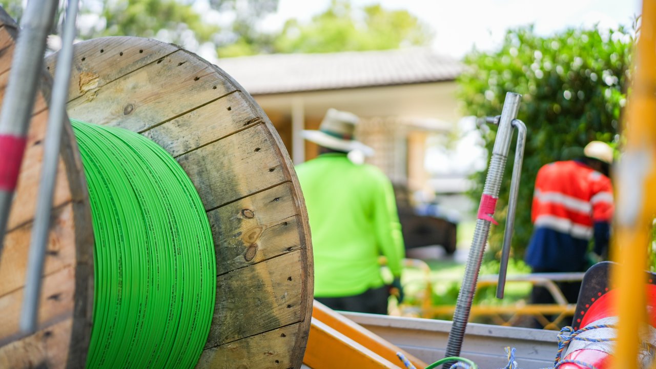 Close-up of a wooden reel holding bright green conduit, with blurred workers and equipment in a residential setting behind.