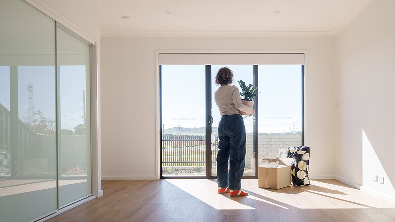 A woman stands looking out glass doors in an empty room. She holds a potplant and there is a box and bags on the floor beside her.