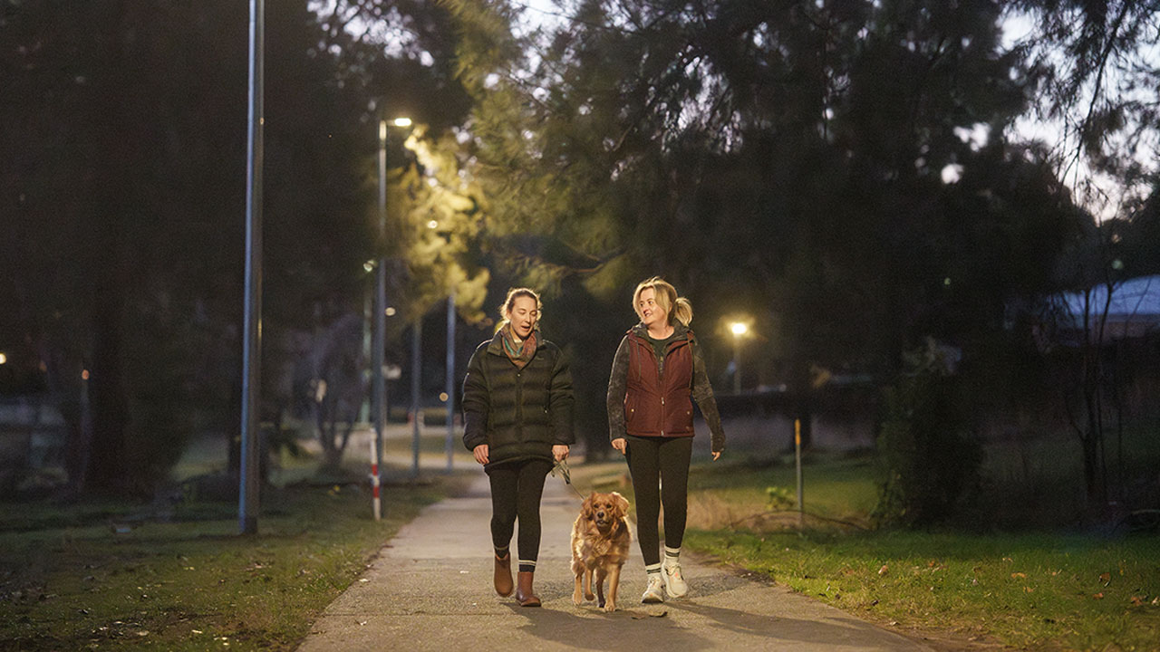 Two women walk a dog along an urban path at dusk. The path is lit by street lights running alongside.