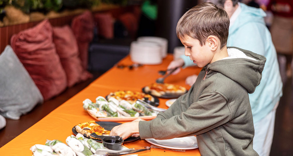 A young boy in a hooded sweatshirt reaches for food from plates of rice paper rolls and pizza on a table. Another person serves themselves in the background.