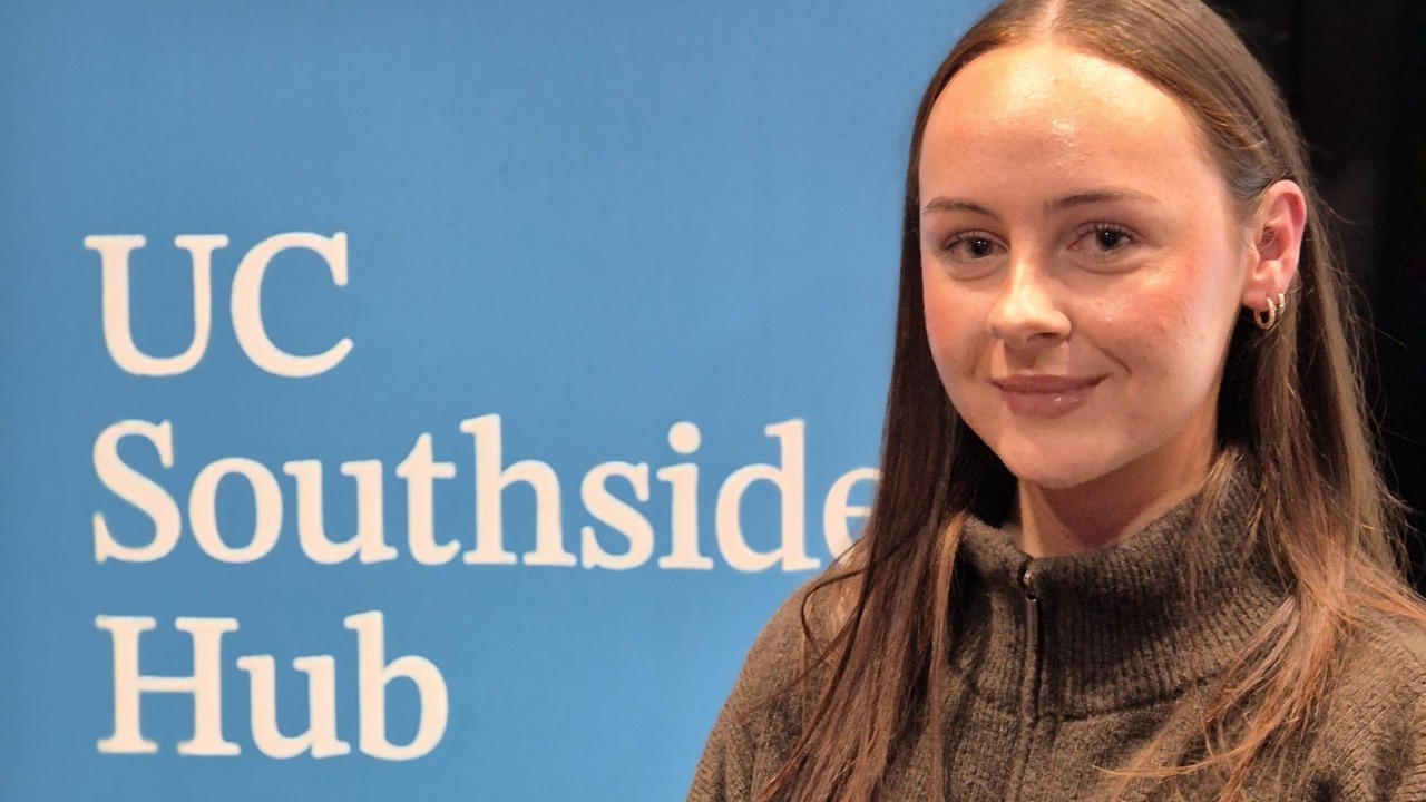 A young woman with long brown hair wearing a brown zip-neck knit sweater stands in front of a blue banner displaying the text “UC Southside Hub.”