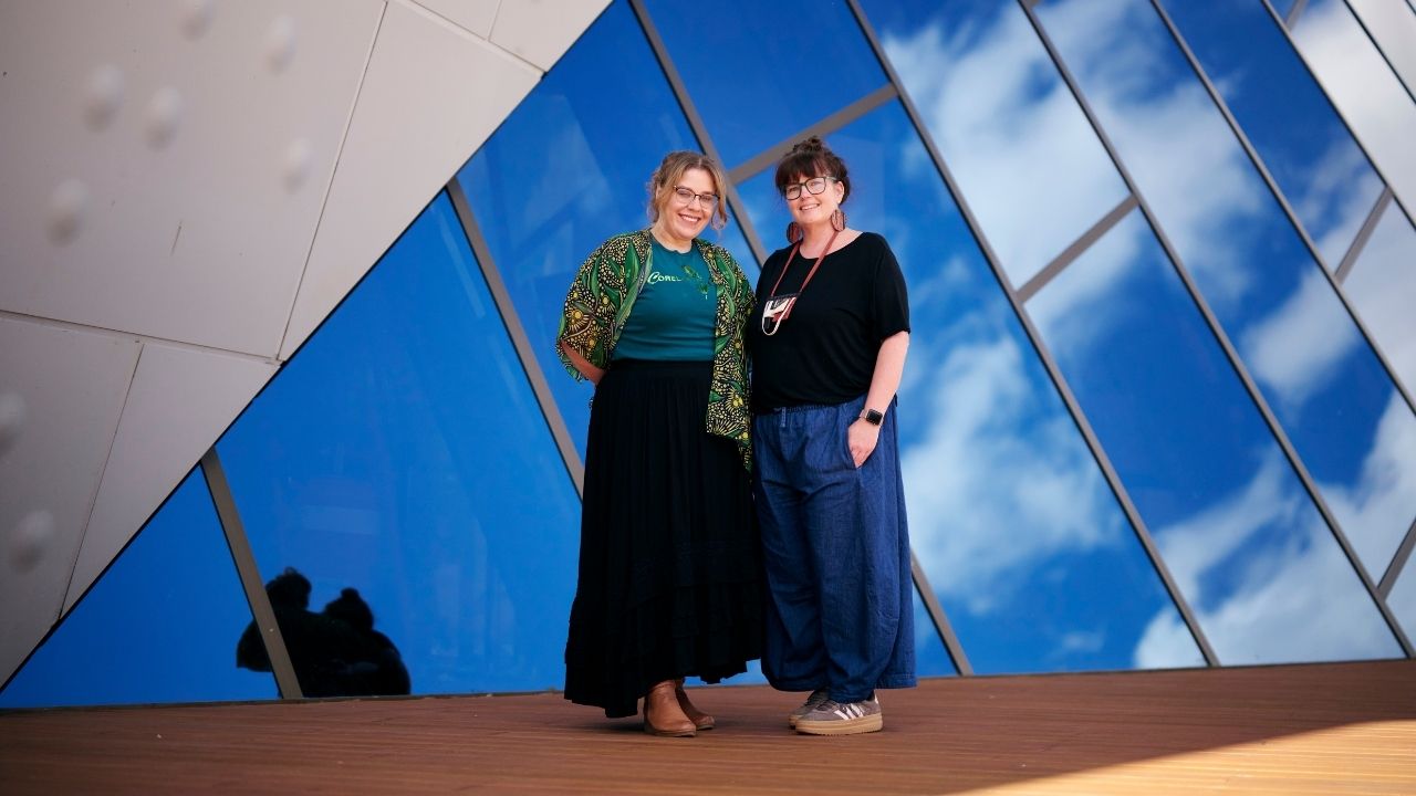 Two people stand side by side on a timber deck in front of a modern building with large blue-tinted, angular glass panels reflecting clouds in the sky.