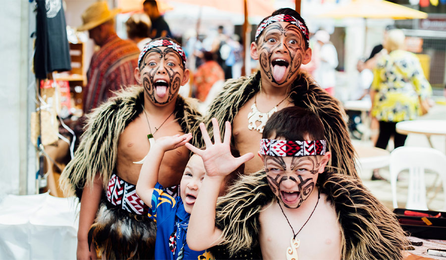 Група момчета, облечени като островитяни с белези по лицето, маха и изплези език 