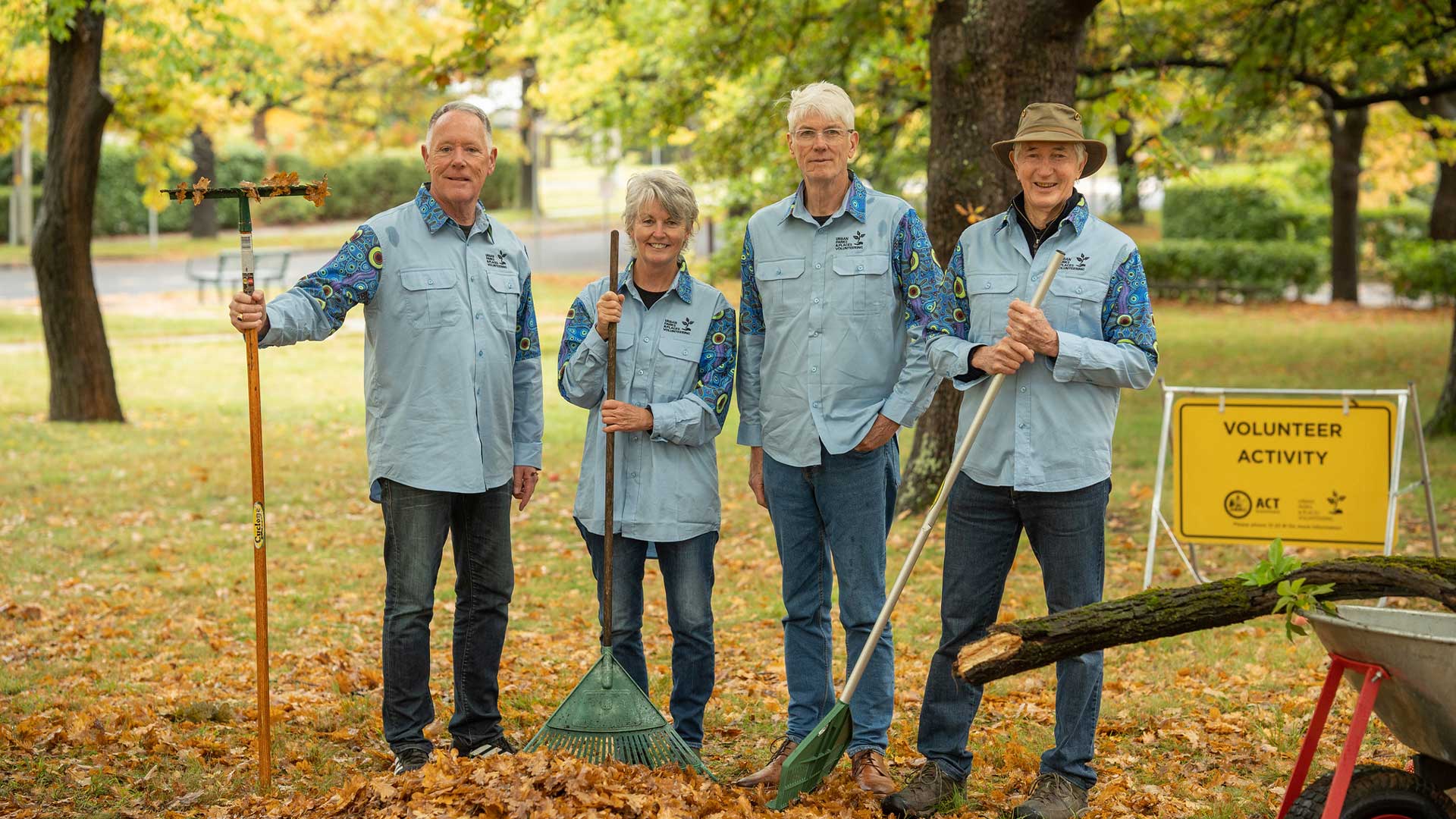 A group of four people stand with rakes beneath trees. There are leaves on the ground and a sign behind them that reads 