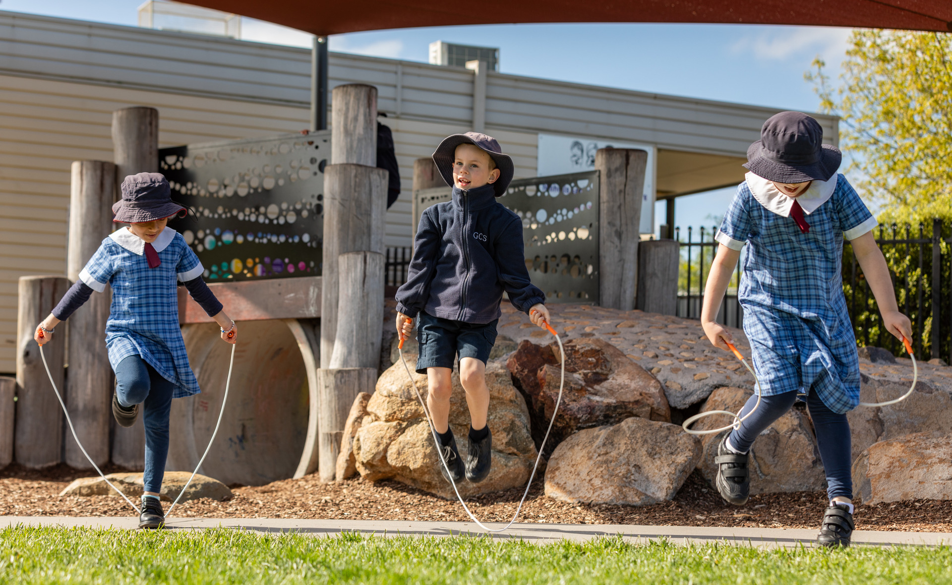 Three children in school uniform skip in front of a playground.