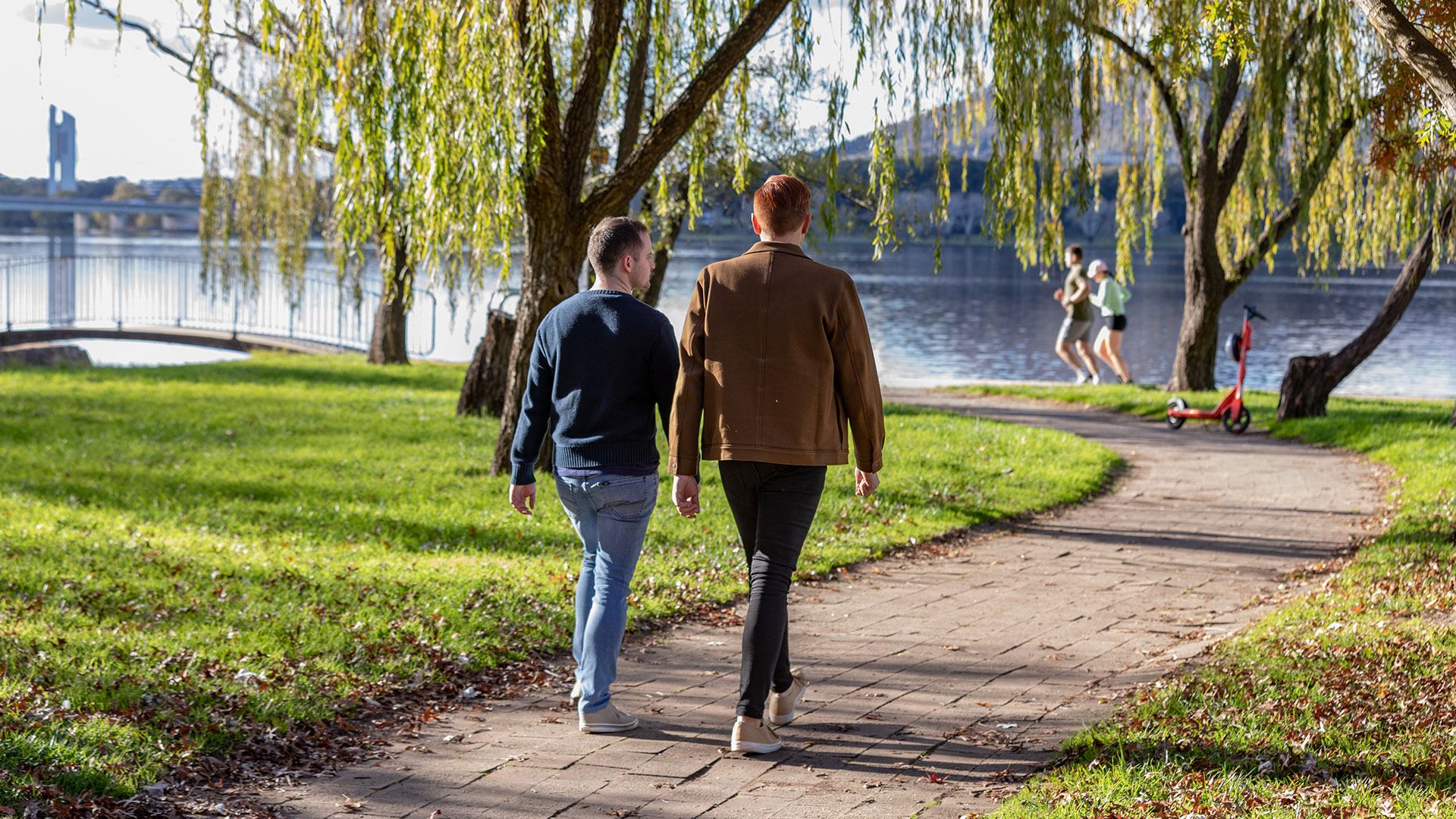 Two men walk by a lake. They are photographed from behind.