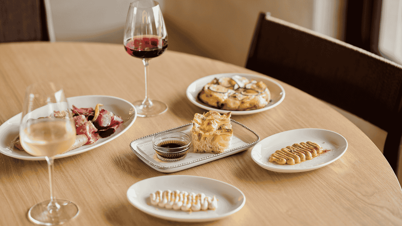Dining table set with multiple dishes, accompanied by glasses of wine, in a well-lit room.