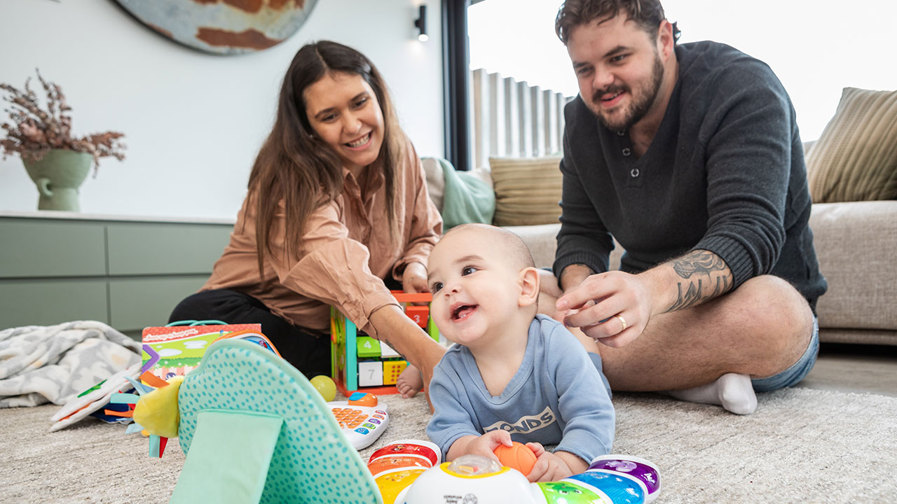 A smiling baby lies on the carpet surrounded by colourful toys, while two adults sit beside them, laughing and playing together in a bright, modern living room.
