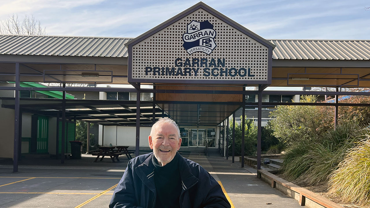 An older man in a wheelchair smiles at the camera. He sits at the entry of a primary school building.