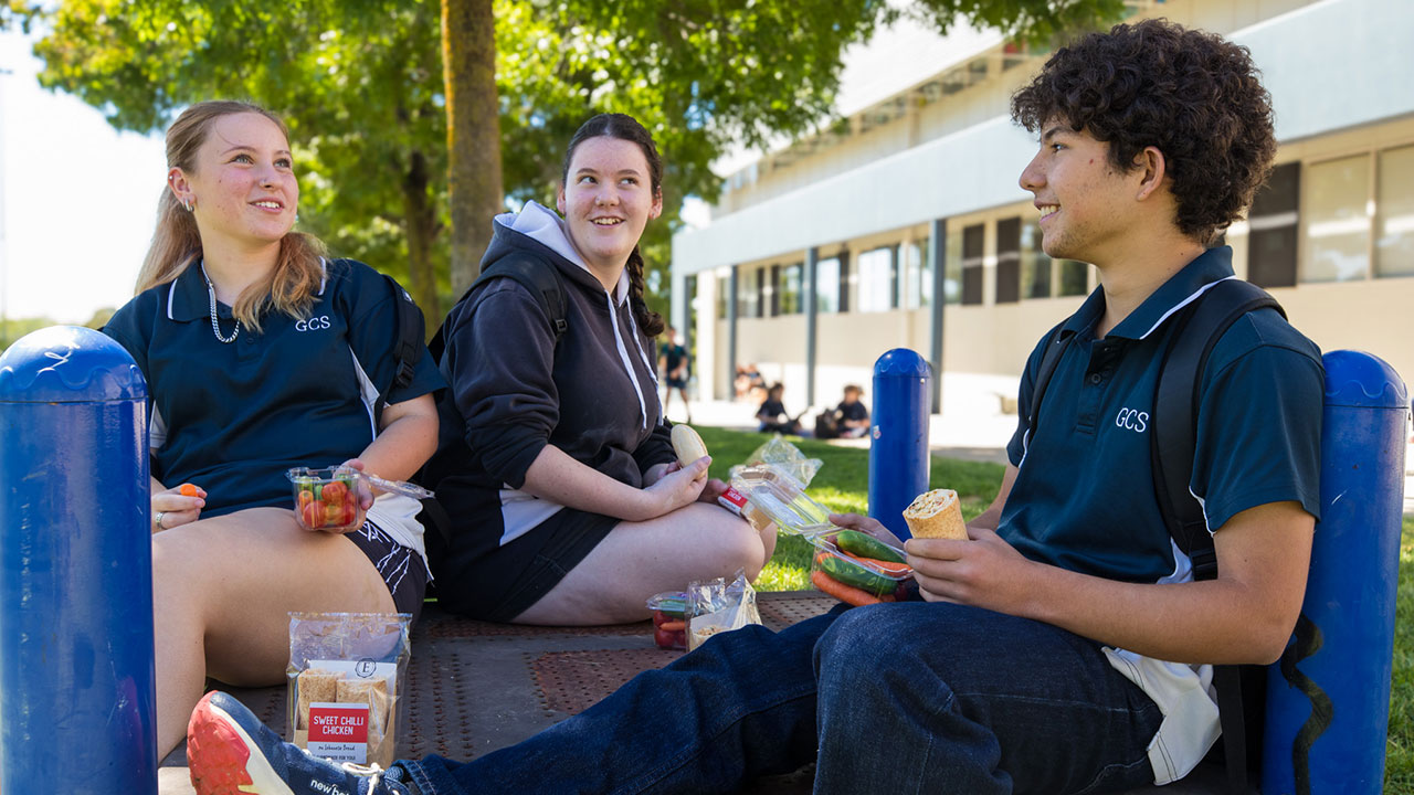 Three teenagers in school uniforms sit outdoors eating lunch.