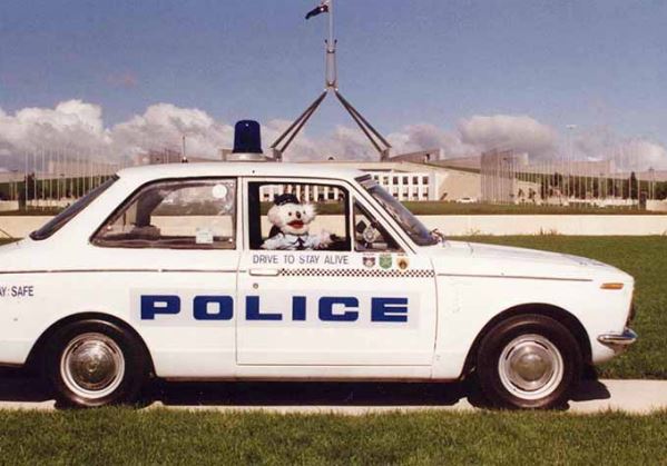 A koala puppet in a police uniform is sitting in a car in front of Australian Parliament House. 