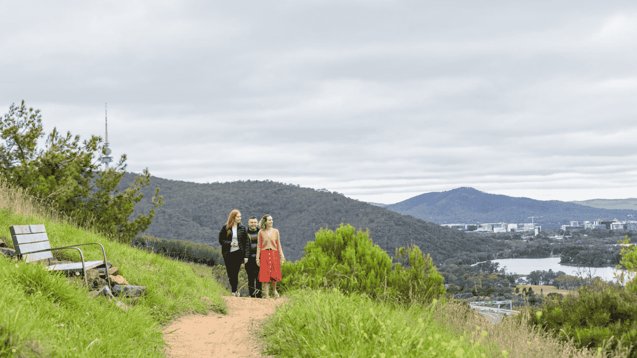 Three people walk on a path surrounded by bush and mountains in the background.