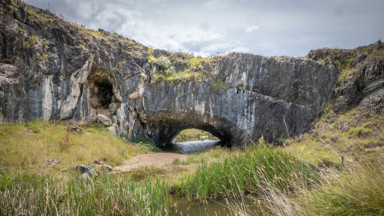 Rocky stone outcrop with a natural arch spanning a narrow stream, surrounded by grassy vegetation and reeds, under a cloudy sky.