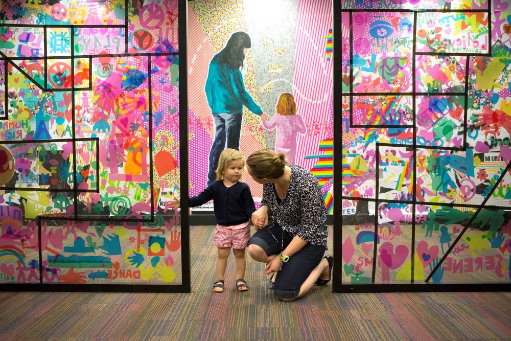 A small child stands beside a crouching adult in front of colorful glass panels filled with bright, playful designs. Behind them, a mural shows two figures holding hands and walking up rainbow-striped stairs.