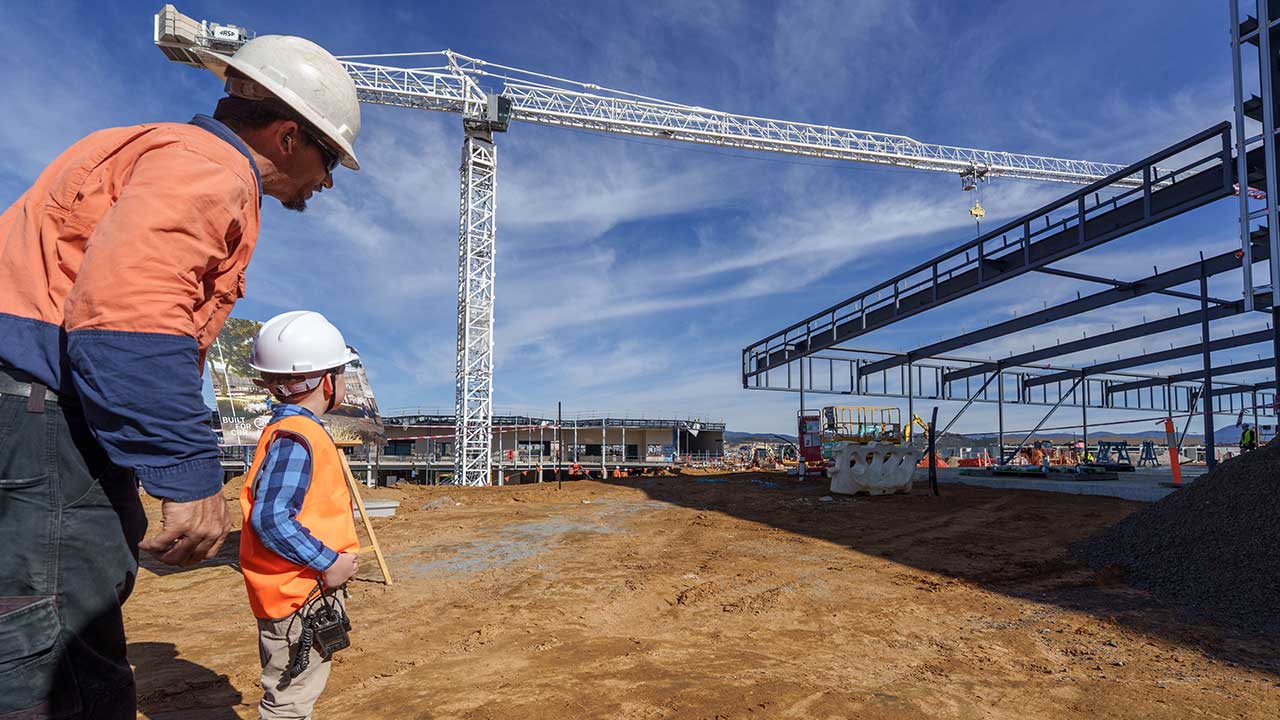 An adult man and a young boy wearing hard hats and high-visibility clothing stand at a construction site beside a tower crane and steel building frame under construction.