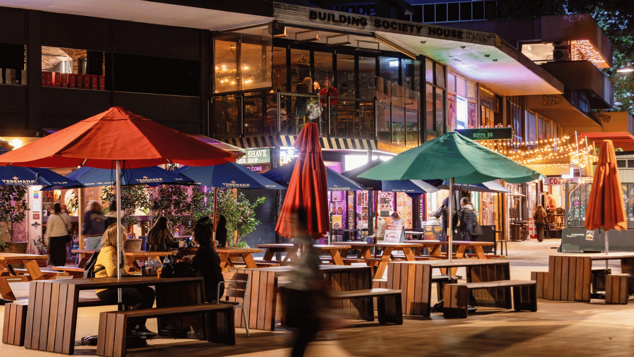 Outdoor dining area in a lively urban entertainment precinct at night, with wooden picnic-style tables, large red and green umbrellas, illuminated shopfronts and restaurants, and people walking and dining.