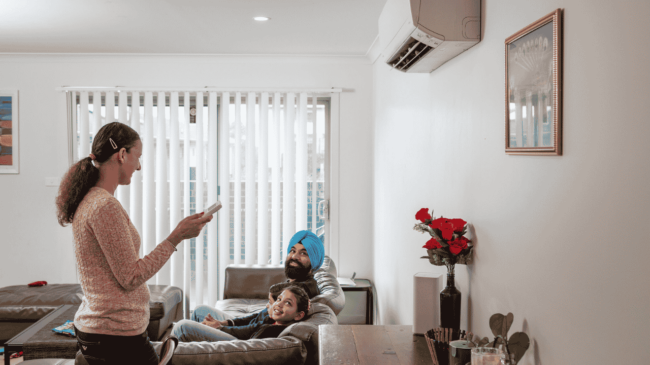 A person stands in a bright living room holding a remote control and looking toward a wall-mounted air conditioner. Two people are seated on a leather sofa nearby.