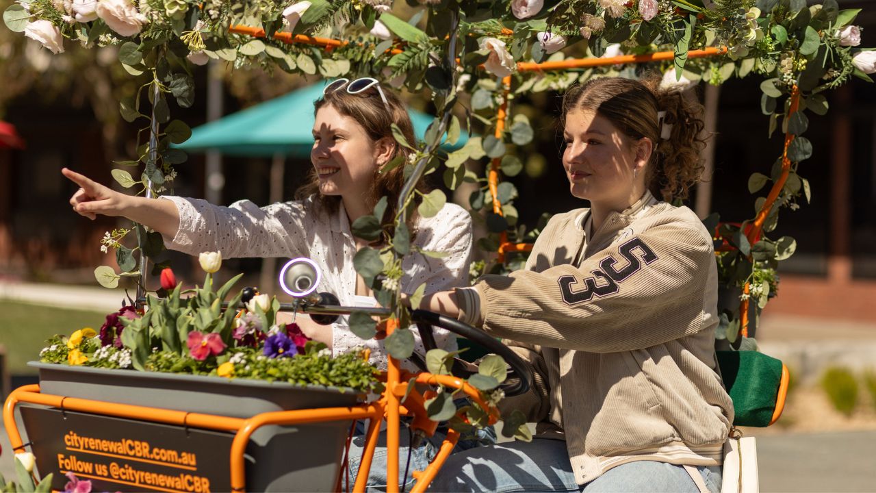 Two girls sit upon an orange tandem bike decorated with flowers