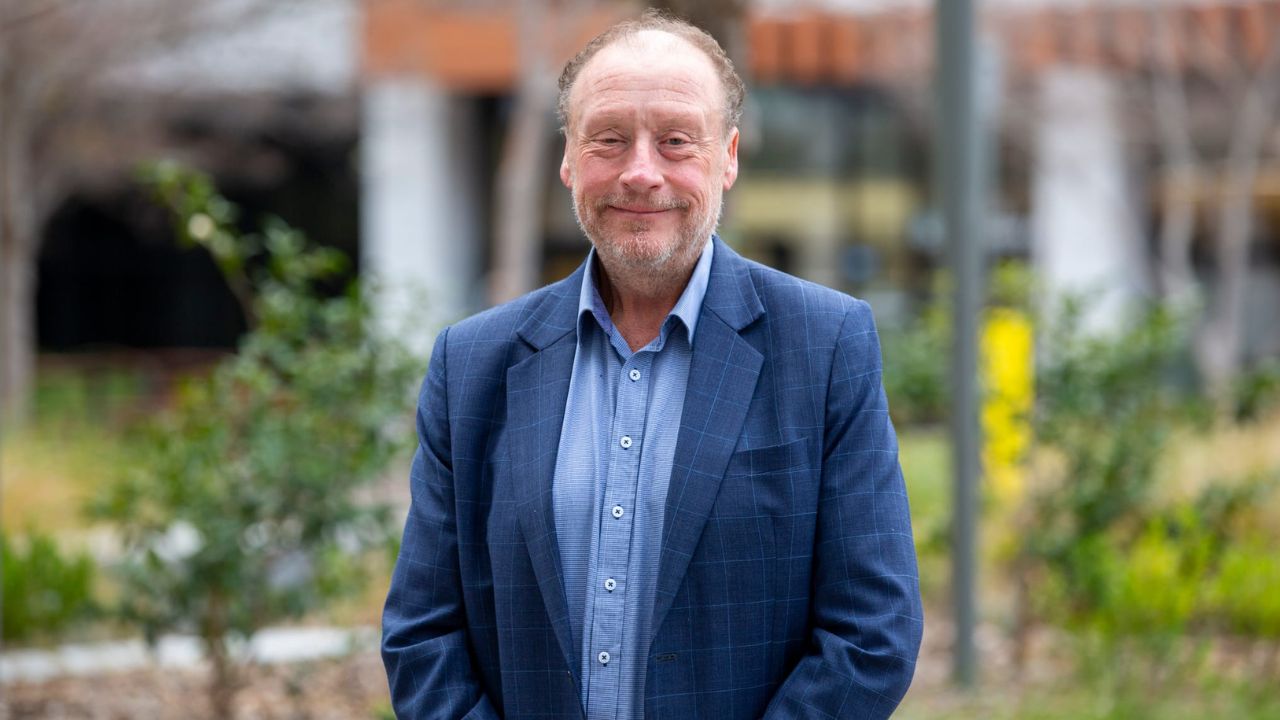Man stands wearing blue jacket in front of garden. He is smiling. 
