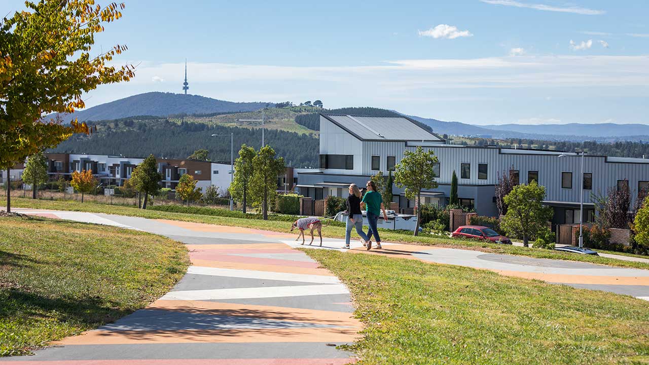 Two women walk a dog along a colourful path overlooking housing. Telstra Tower is in the background.