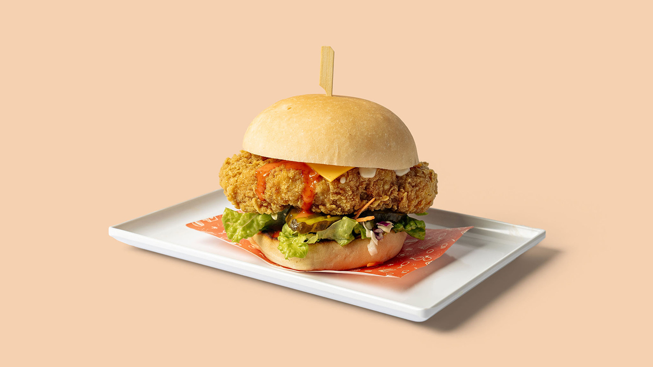 A chicken burger with cheese, lettuce. pickles and sauce in a white bun, set against a peach-coloured background.