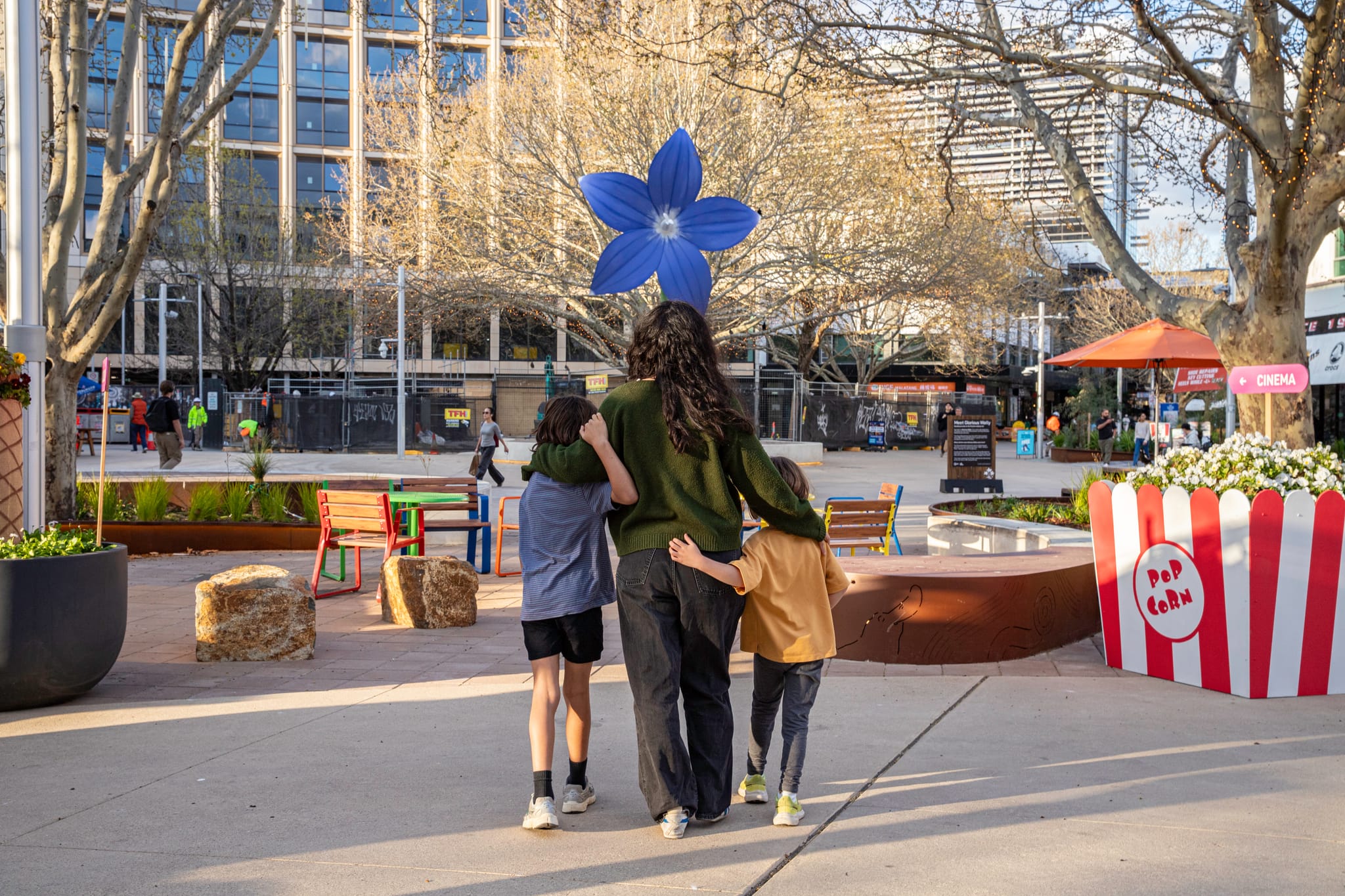 A parent walks into Garema Place with her two kids where a giant flower is located with colour trees and furniture