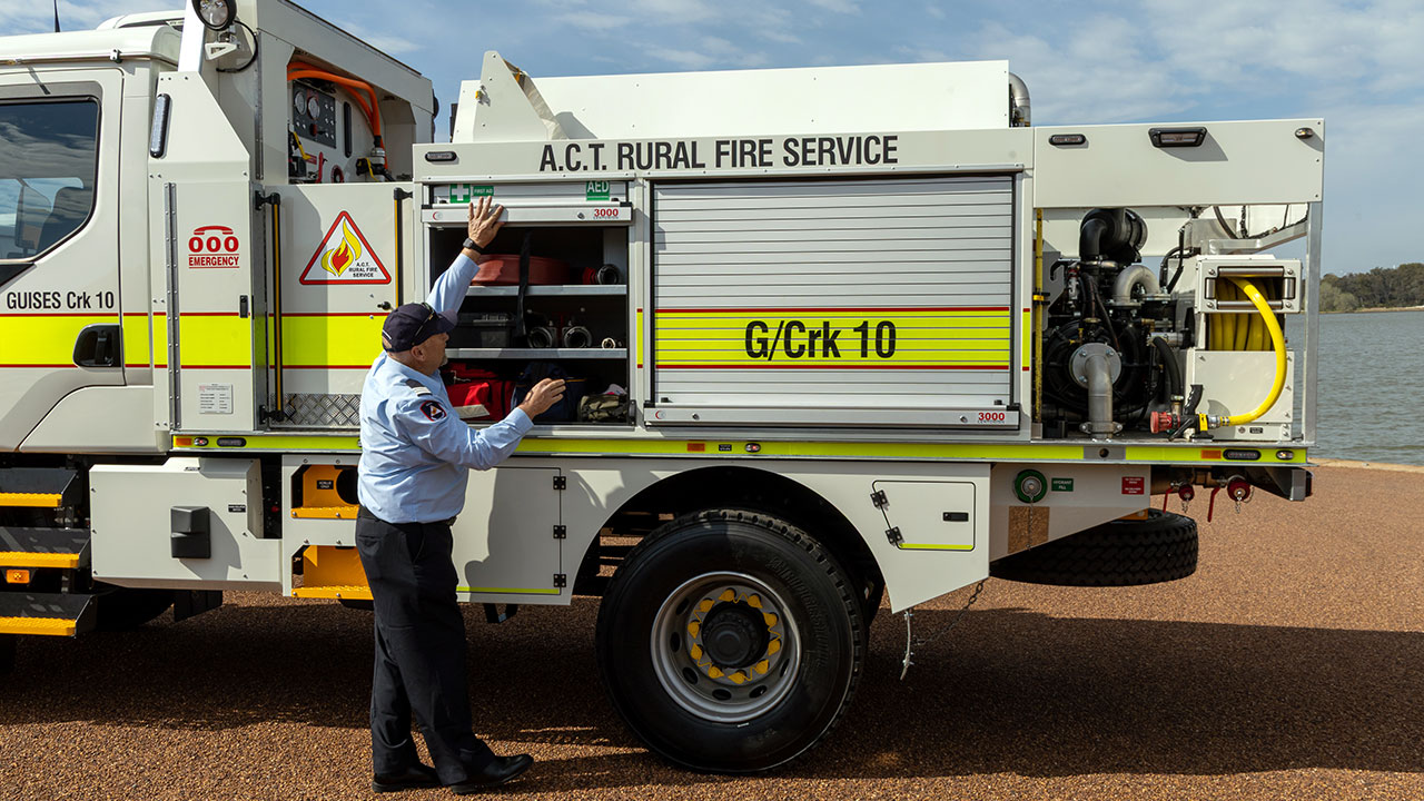 A man in uniform stands beside a fire truck, showing at the equipment inside.