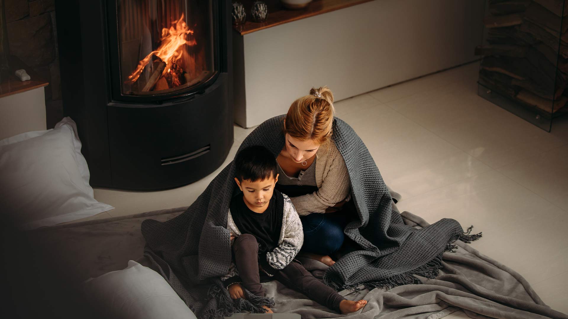 Mother and child sit by wood heater with blanket. 