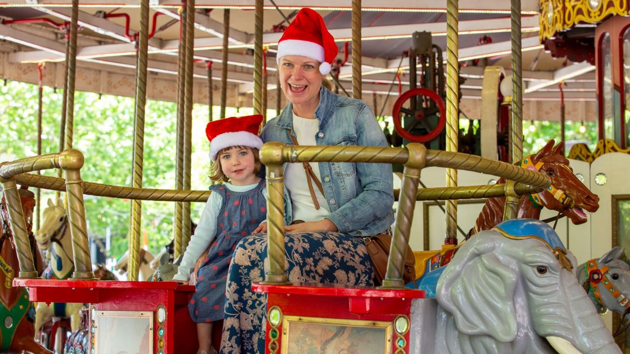 Mother and daughter wearing santa hats ride on merry-go-round.