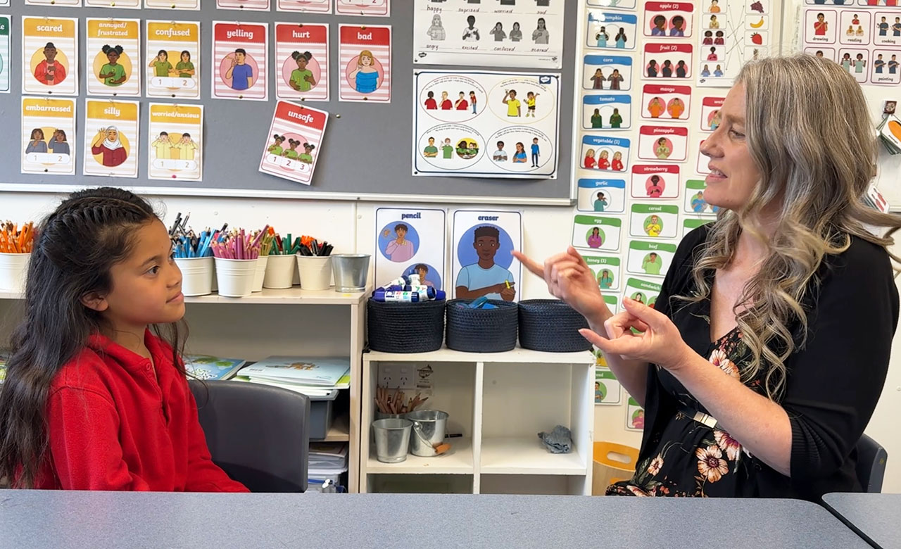 A young girl in a school uniform sits attentively as an adult woman uses sign language in a classroom.