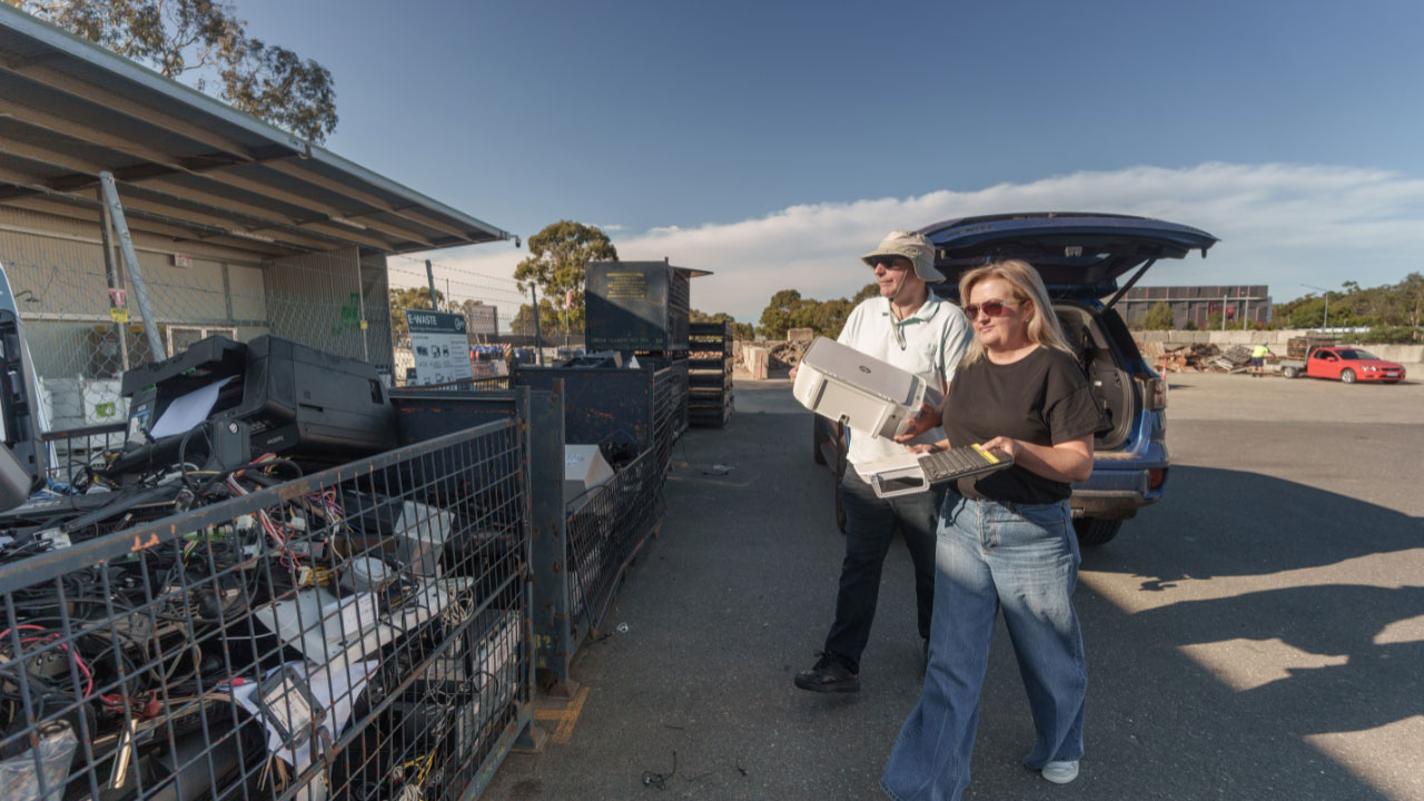 A man and woman carry a keyboard and printer towards a cage filled with disposed electrical waste