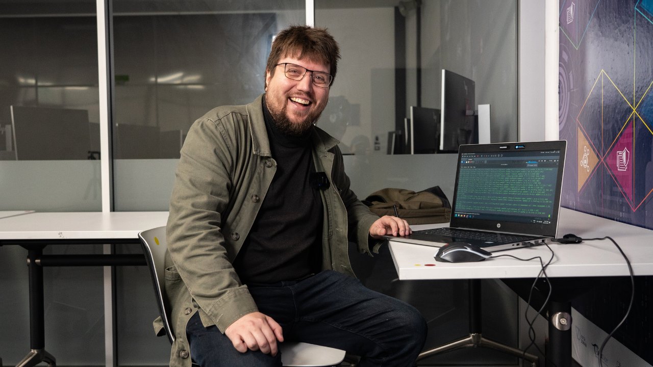 A bearded man sits in front of a laptop showing code. He smiles.