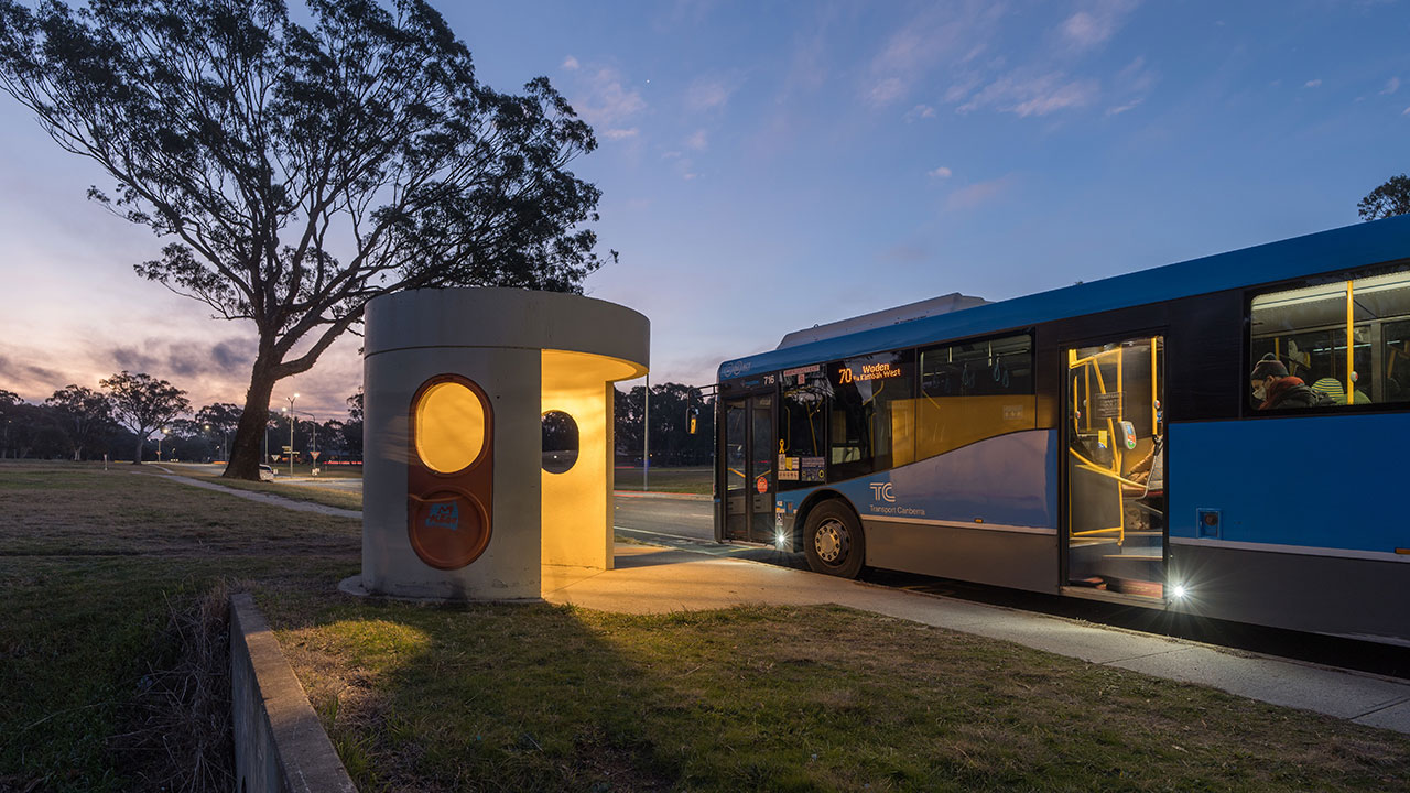 A blue bus stopped beside an ACT bus shelter at dusk. The bus shelter is lit from the inside.