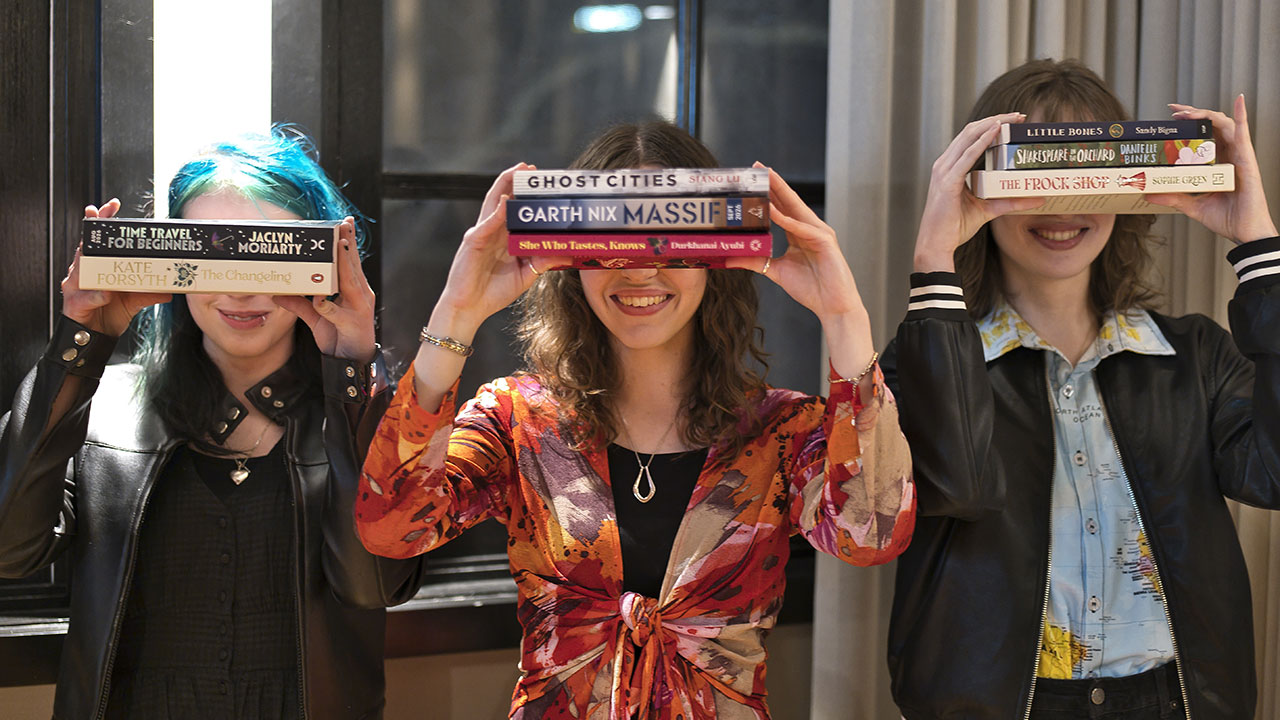 Three people stand next to each other holding up piles of books that obscure their faces.