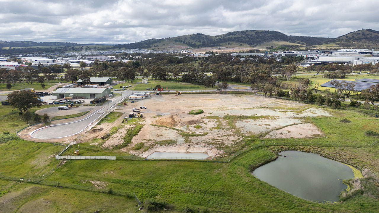 An overhead image showing an area of bare dirt surrounded by grass, bush, and housing in the distance.