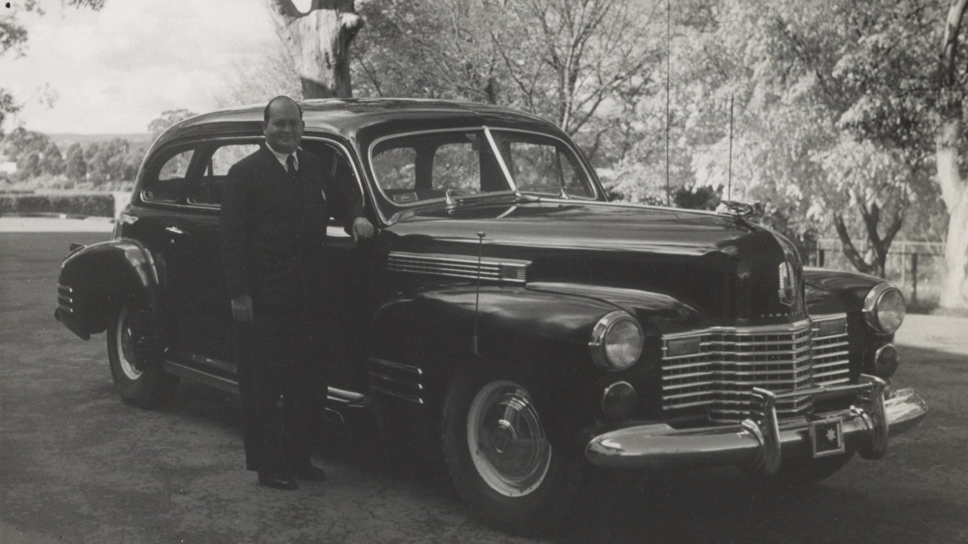 A black and white photo showing a man standing by a dark old-style car.