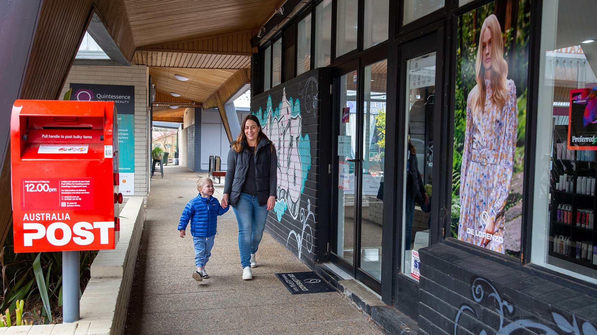 A woman and young child walk along a local shopping area. There is an Australia Post box beside them.
