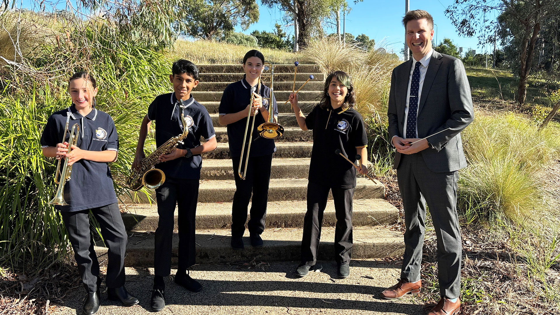 An adult man and four children stand on some steps outside. Each of the children is holding a musical instrument. They are all smiling.