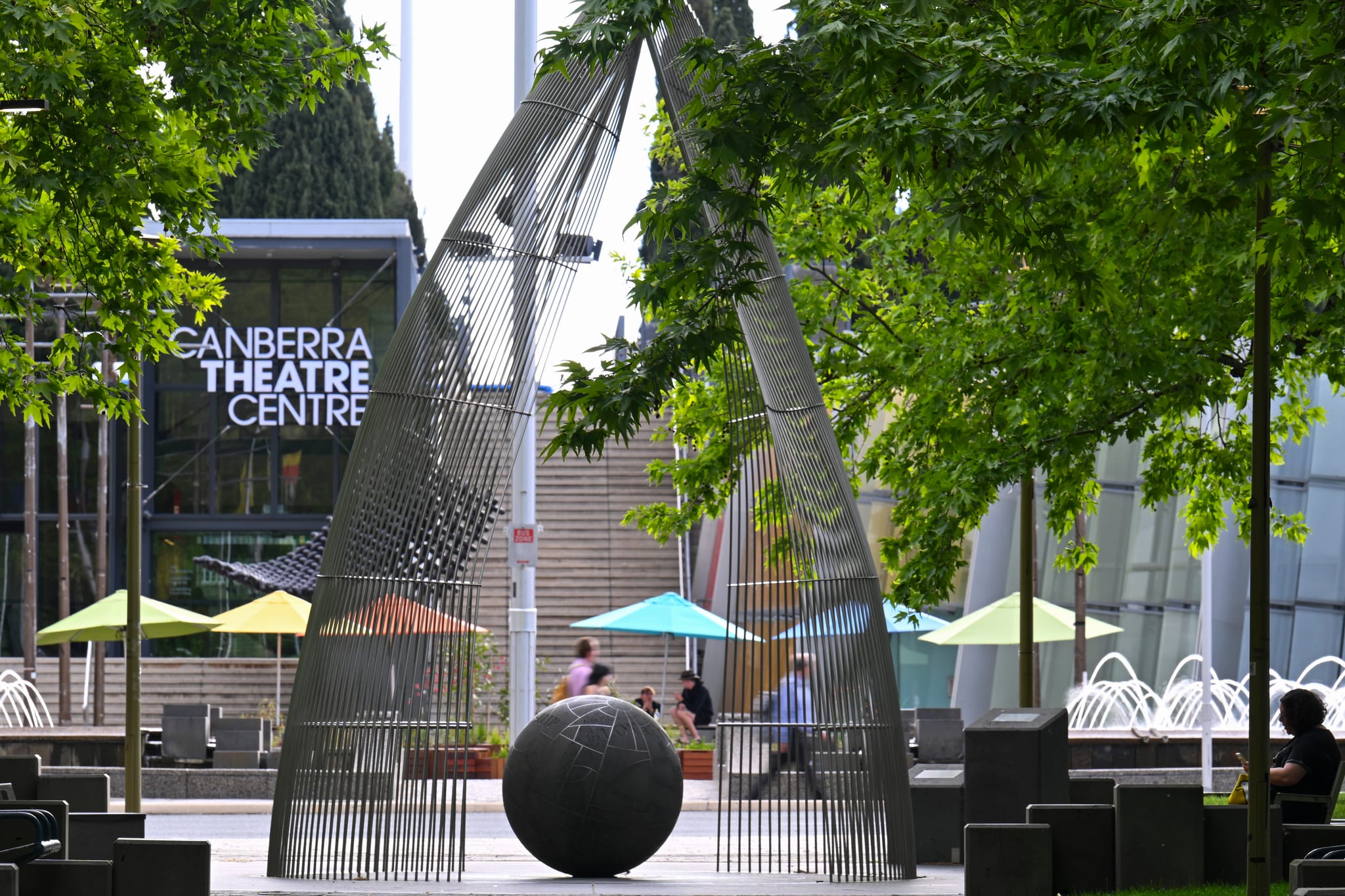 A view of Civic square looking across Ainslie Place with colourful umbrellas and tables, a sculpture and the Canberra Theatre Centre, fountain and people in the background