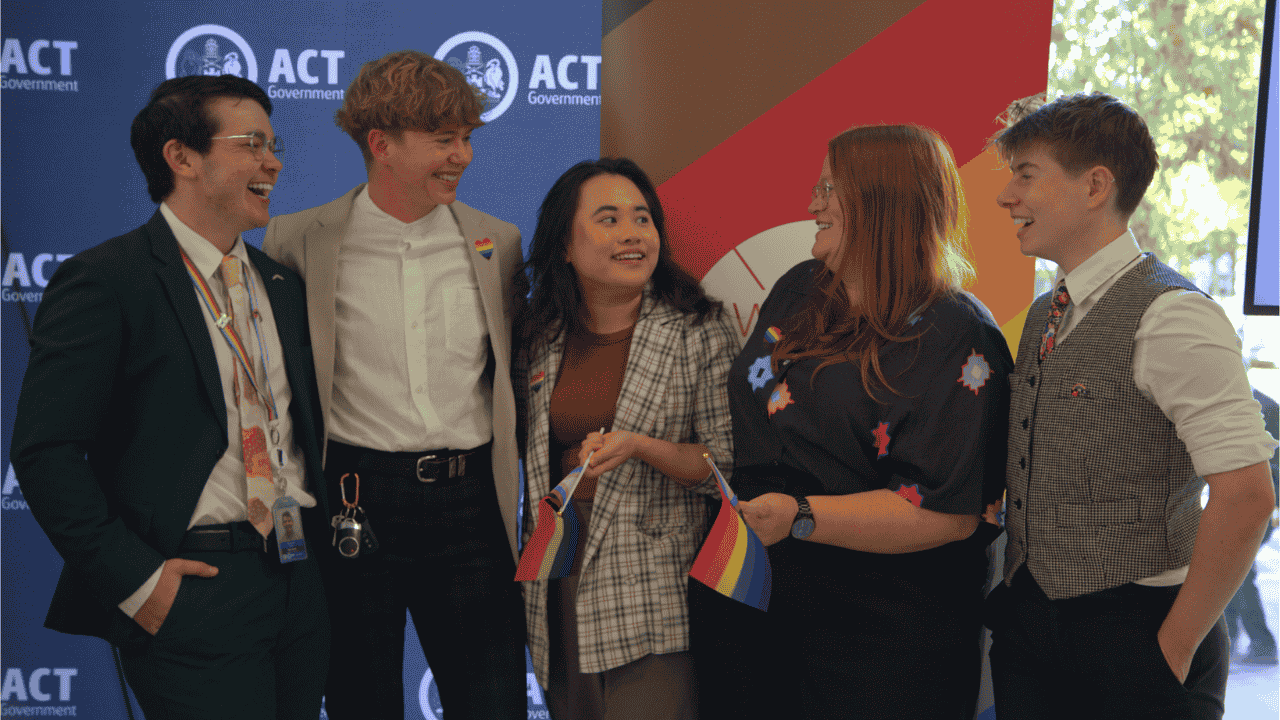 A group of people laugh together with a rainbow banner in the background and the logo of ACT Government.