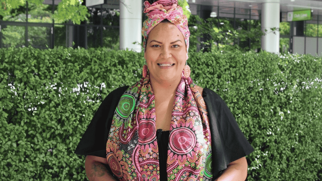 A woman wearing a patterned scarf stands in front of green bushes.