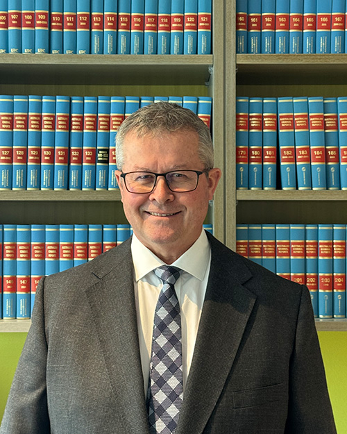 A man with grey hair and glasses smiles while standing in front of a bookcase of law books