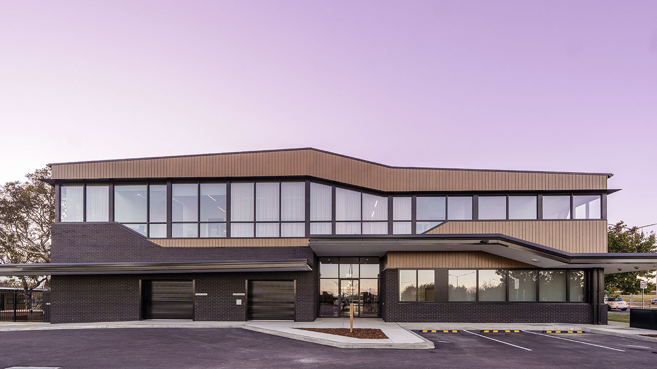 Modern two-storey community building with large glass windows and dark brick at ground level and light timber-style cladding on the upper level, set in a paved car park with marked spaces in front.