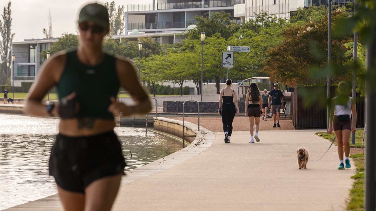 A person running on a wide footpath beside a lake with people walking in the background.