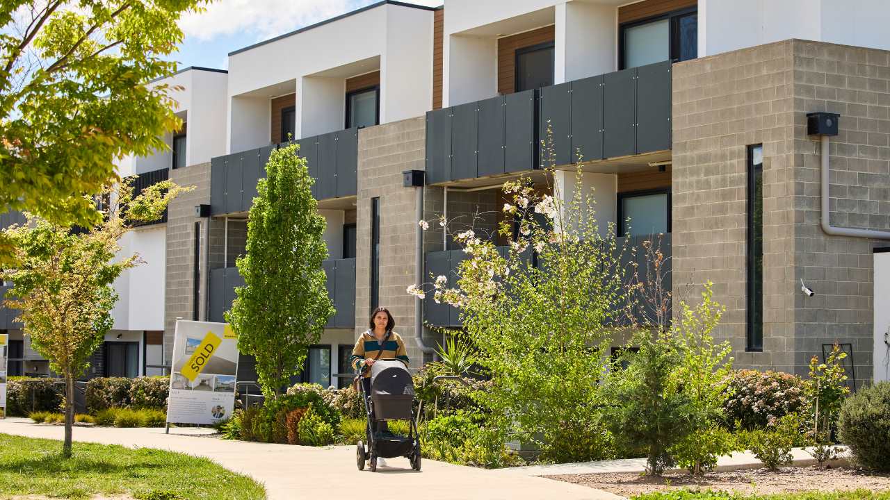A young woman walks with a pram along a street of modern townhouses. There is a Sold sign out the front of one of them.