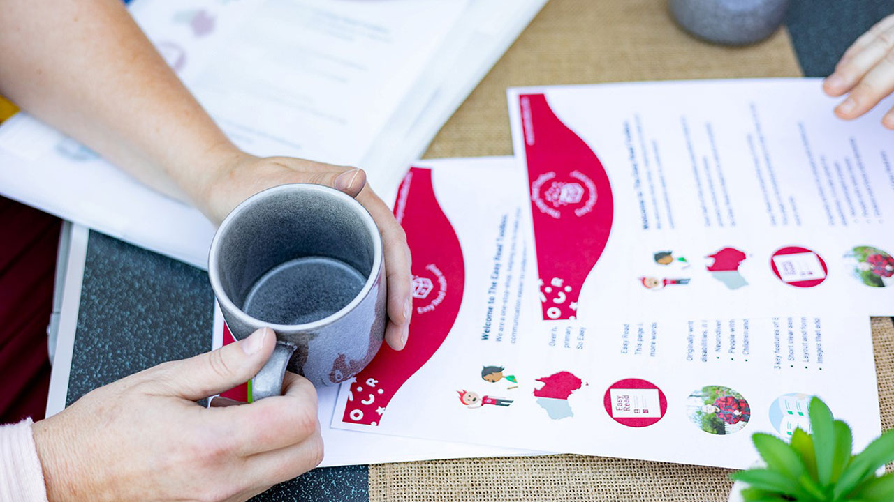 Hands holding a mug of tea above papers laid out on a table.