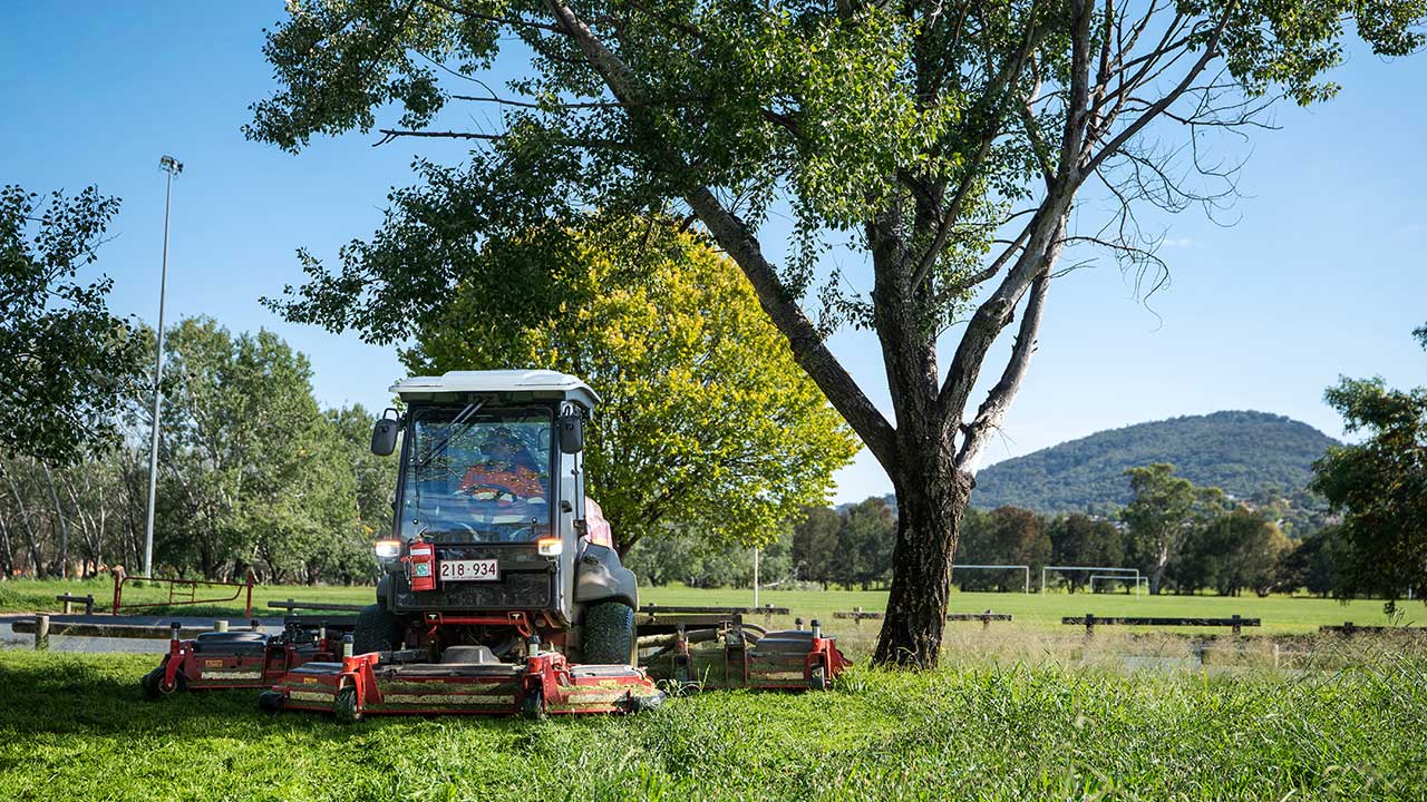 A man dressed in high-visibility clothing drives a large ride-on mower across long grass with a sporting field and mountains in the background.