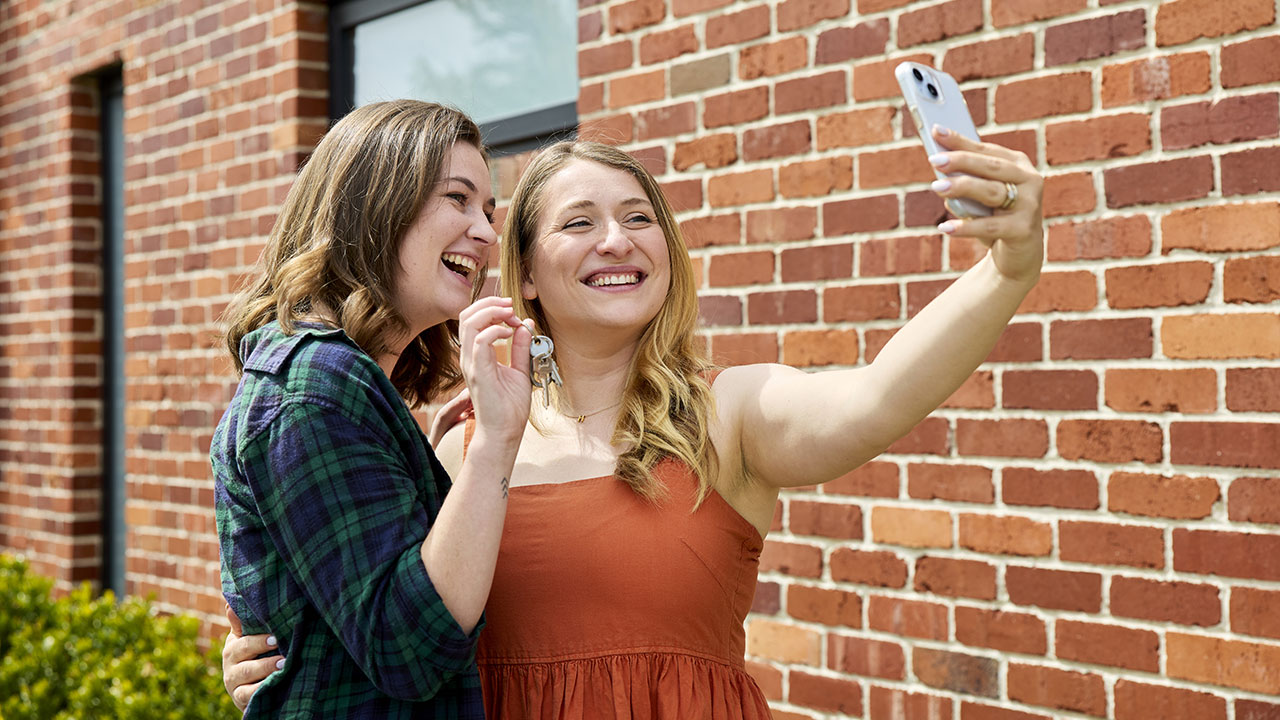 Two young women stand together taking a selfie. One holds up a set of keys.