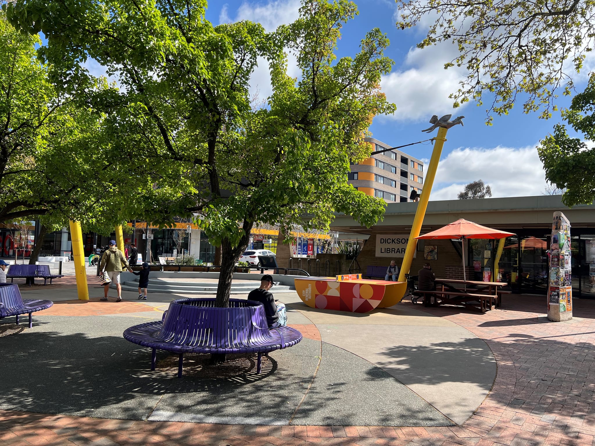 Image shows the current state of Dickson Shops with colourful purple bench seating, shade sail, orange umbrella and City Renewal branded ping pong table. Someone enjoys reading a book in the shade of a tree.
